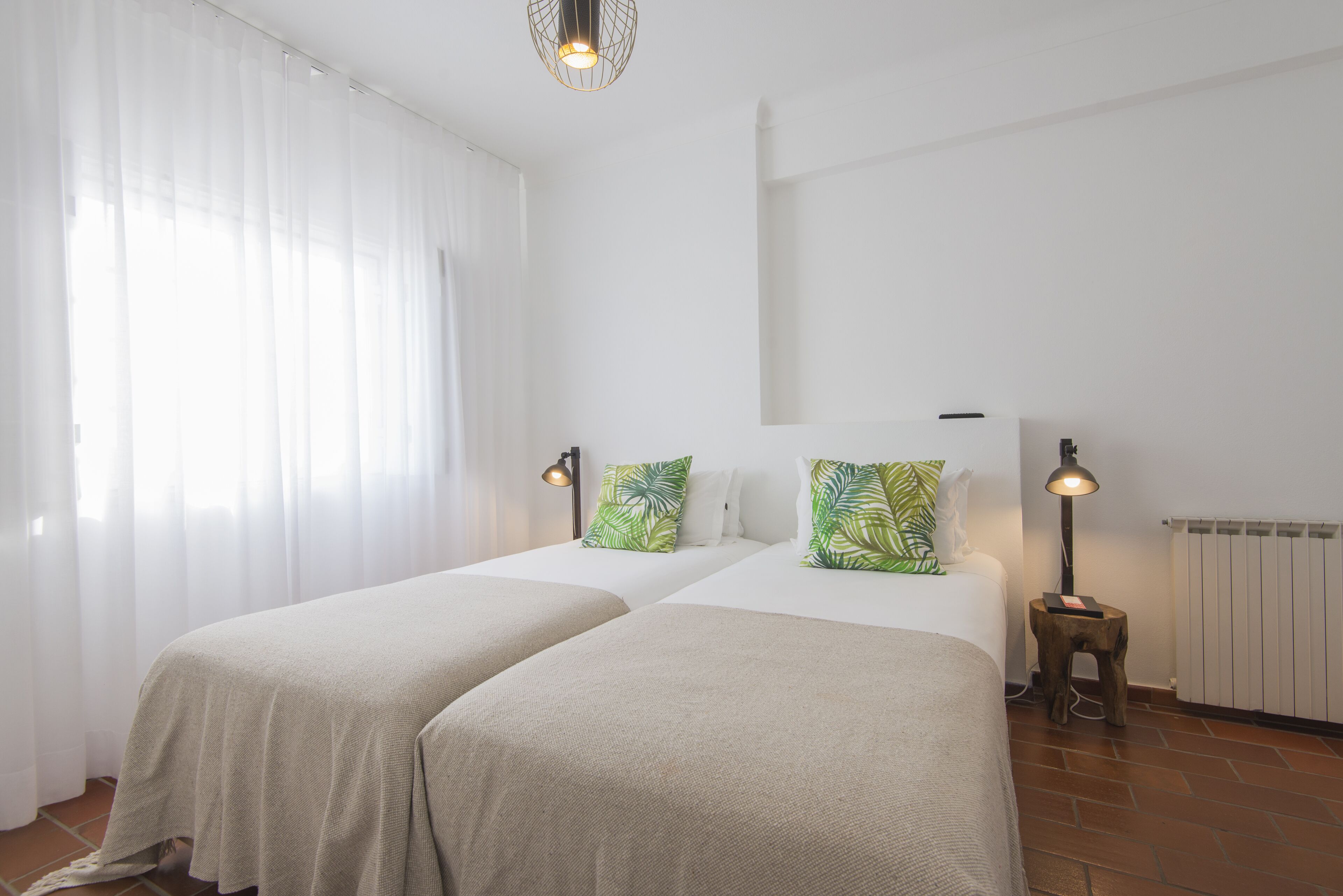 A room with two beds, white walls, a radiator, and a window with sheer curtains.