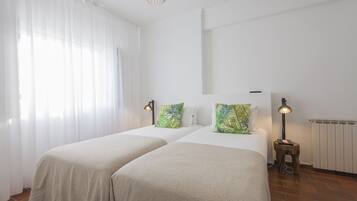 A room with two beds, white walls, a radiator, and a window with sheer curtains.