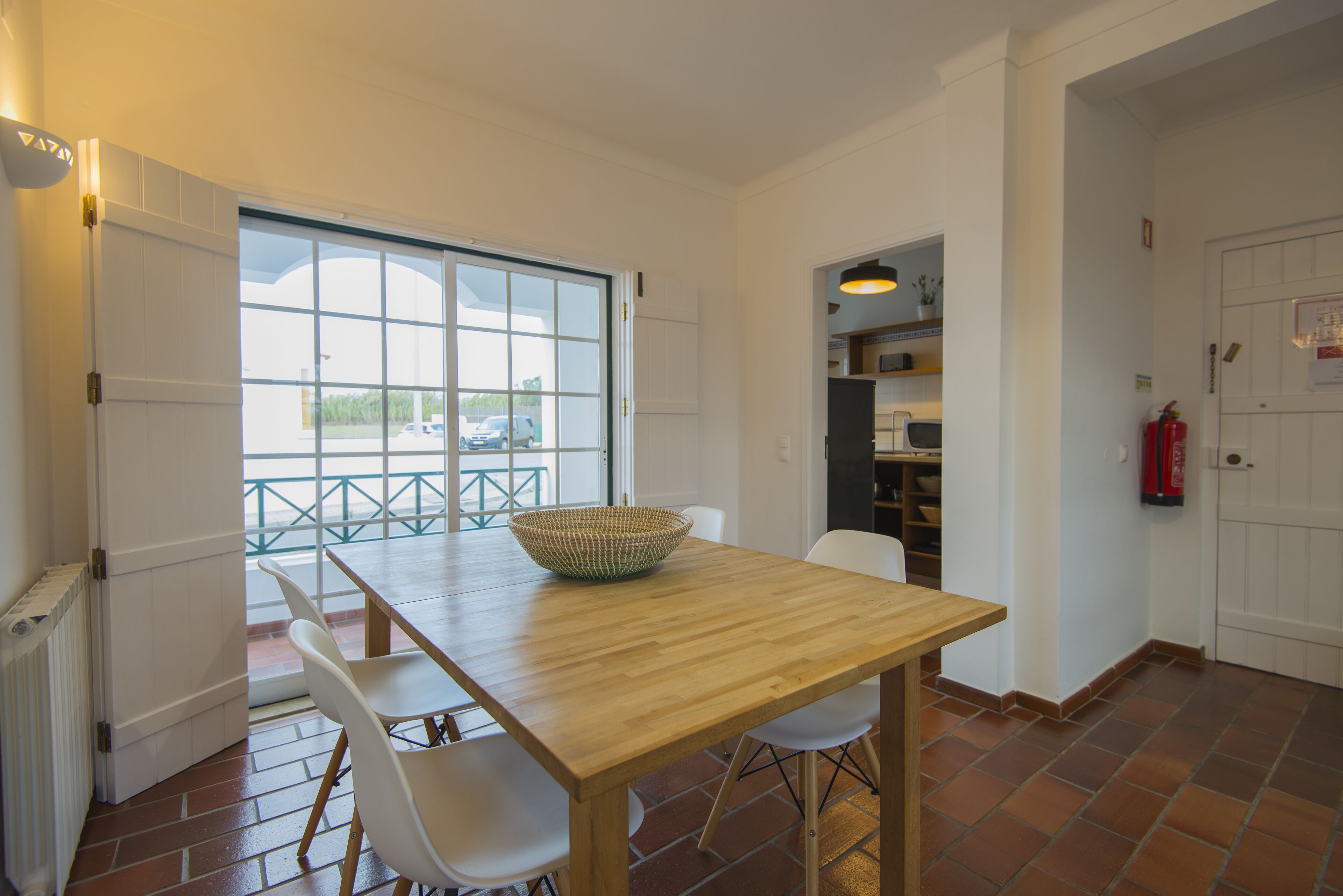 A dining area with a wooden table and white chairs, a large window with a view of the outside, and a kitchen area visible through an open door.