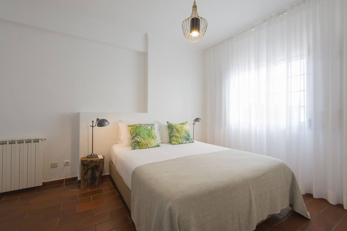 Une chambre à coucher avec un lit, des rideaux blancs, un radiateur et une table de chevet en bois.