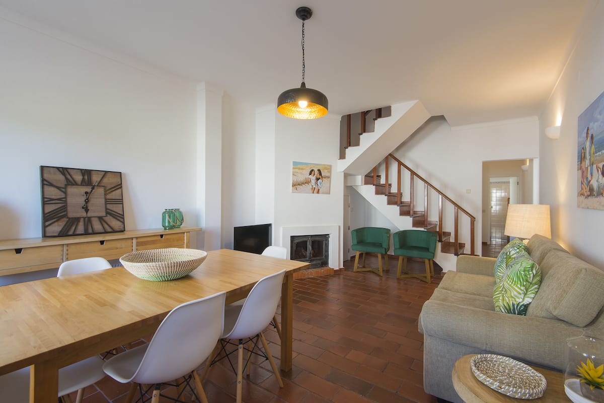 Un vaste espace de vie comprenant une table à manger en bois, des chaises blanches, un canapé vert et un escalier menant à l’étage supérieur.