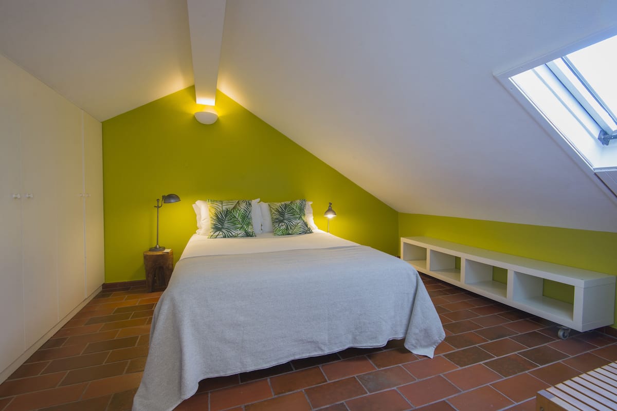 Une chambre avec un plafond en pente, un lit avec une literie blanche, des coussins verts, une table de chevet avec une lampe et un puits de lumière.