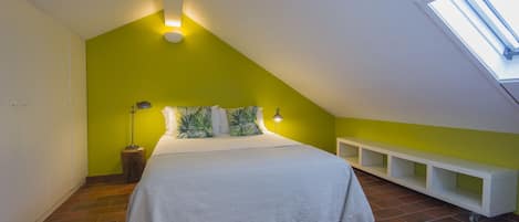 Une chambre avec un plafond en pente, un lit avec une literie blanche, des coussins verts, une table de chevet avec une lampe et un puits de lumière.