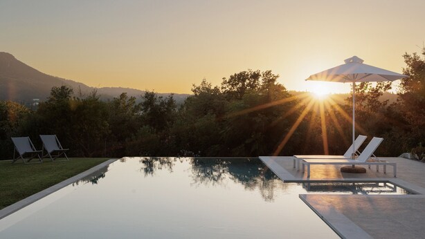Una piscina con sillones de descanso y una sombrilla, rodeada de árboles y una montaña al fondo.
