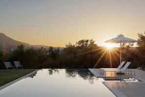 A pool with lounge chairs and an umbrella, surrounded by trees and a mountain in the background.