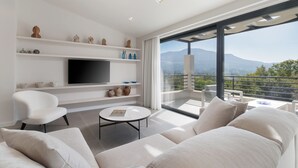 Una sala de estar moderna con televisor de pantalla plana, sofás blancos, una mesa de centro y un balcón con vistas a la montaña.