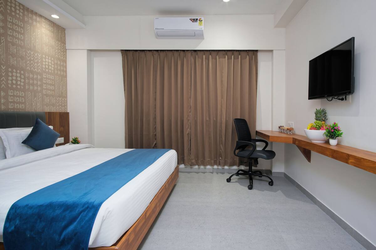 Una habitación de hotel moderna con una cama grande, un escritorio con computadora, un televisor y un ventanal con cortinas.