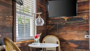 Une pièce aux murs en bois, avec un téléviseur fixé au mur, deux fauteuils en osier, une petite table sur laquelle est posé un vase de fleurs, et une fenêtre avec des stores.