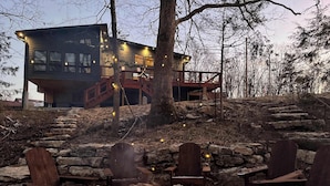 Un área de estar al aire libre rústica con un pozo de fuego de piedra, sillas de madera y una terraza de madera que conduce a un edificio con luces colgantes.