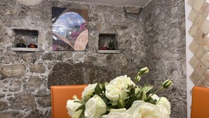 Un mur de pierres sur lequel est accroché un tableau encadré, un vase de fleurs blanches et des chaises orange.