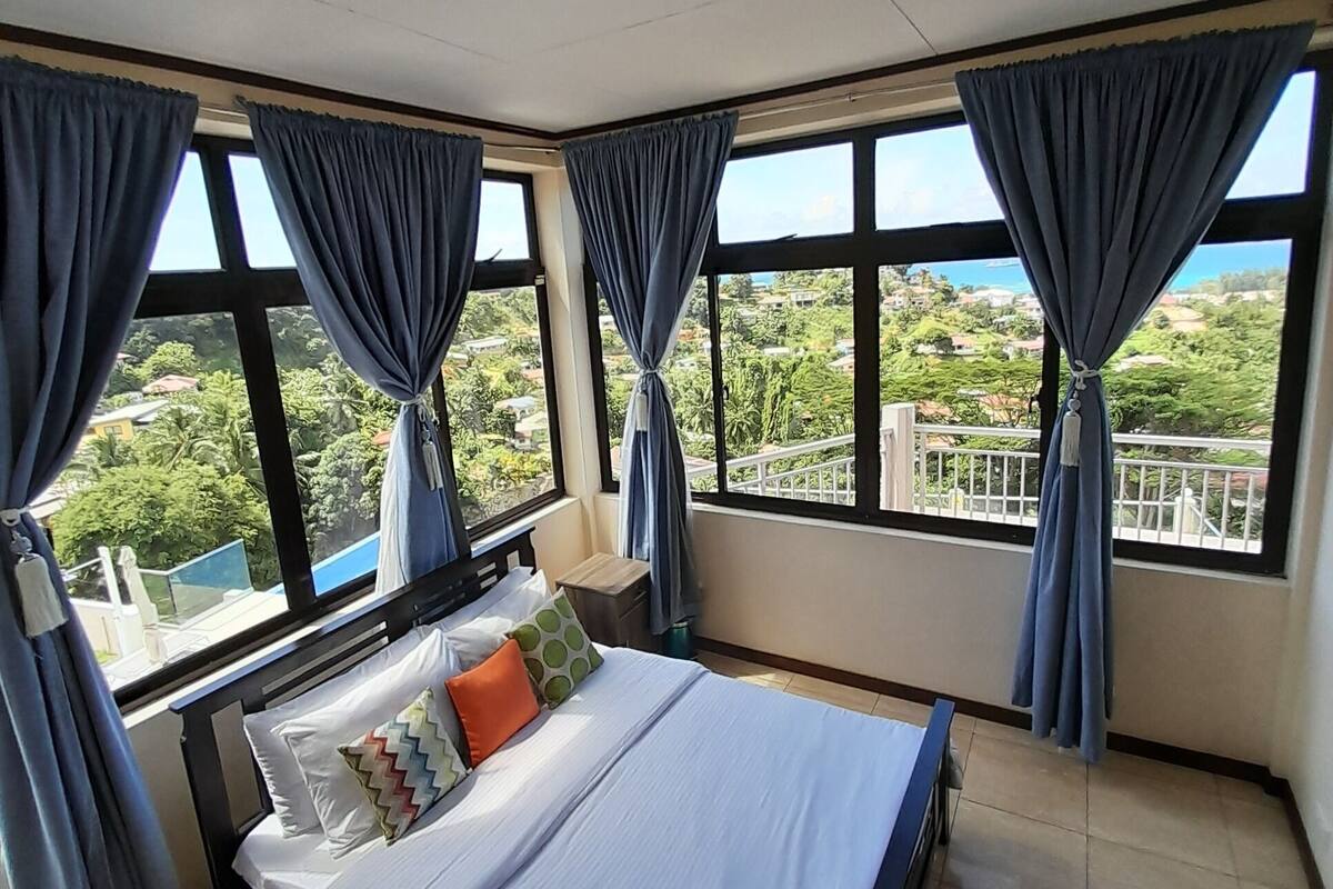 A bedroom with a bed, curtains, and large windows with a view of greenery and buildings.