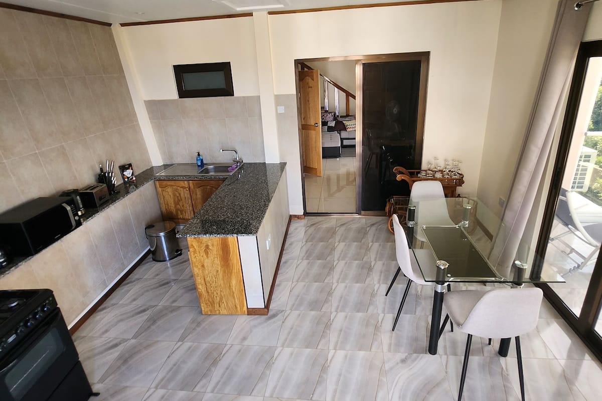 A modern kitchen with a central island, a dining area with a glass table, and a view of the outdoors.