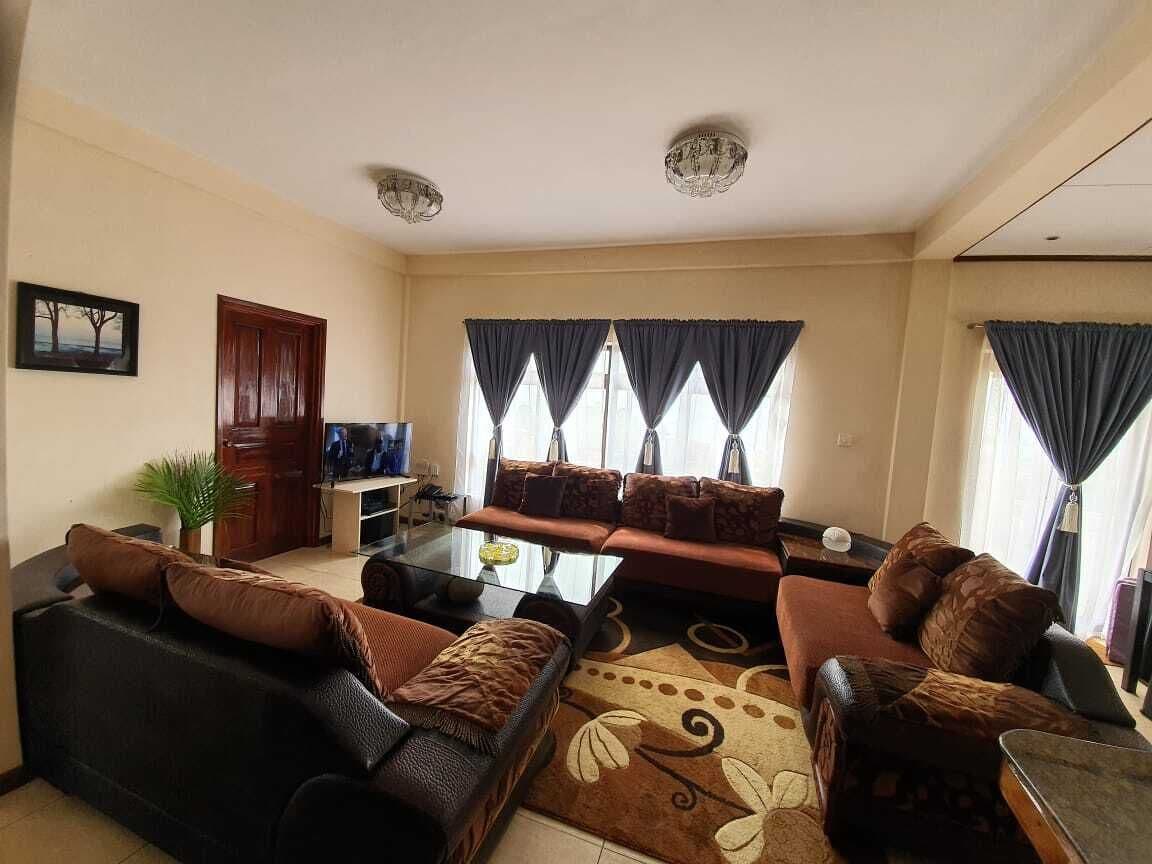 Een woonkamer met een bank, fauteuils, een salontafel en een televisie. Er zijn gordijnen bij de ramen, een deur en een plant.