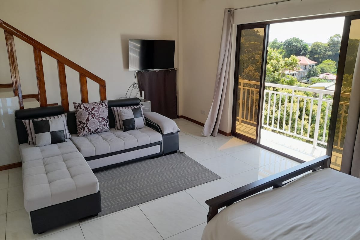 A bedroom with a bed, a sofa, a TV, and a balcony with a view of greenery and houses.