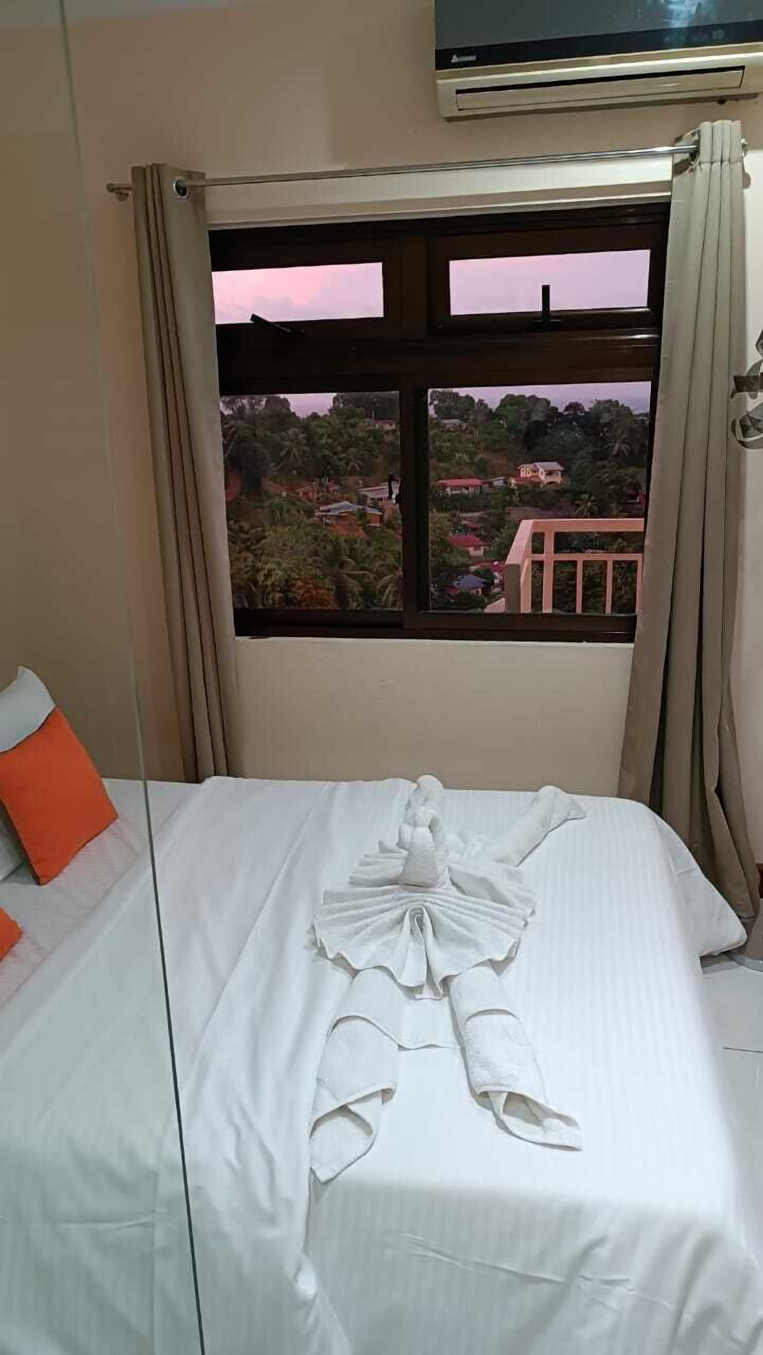 A hotel room with a bed, a window with curtains, and an air conditioning unit above the window.