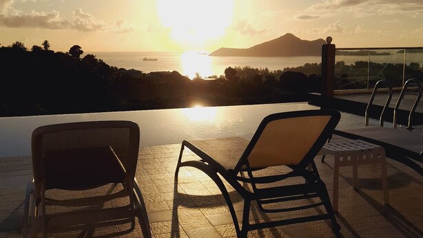 Ein Infinity-Pool mit zwei Liegestühlen, Blick auf einen Sonnenuntergang über dem Meer und einem Berg.