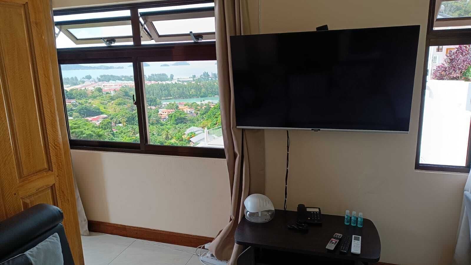 A room with a large window, a flat-screen TV mounted on the wall, a desk with a lamp, a phone, and some bottled water.