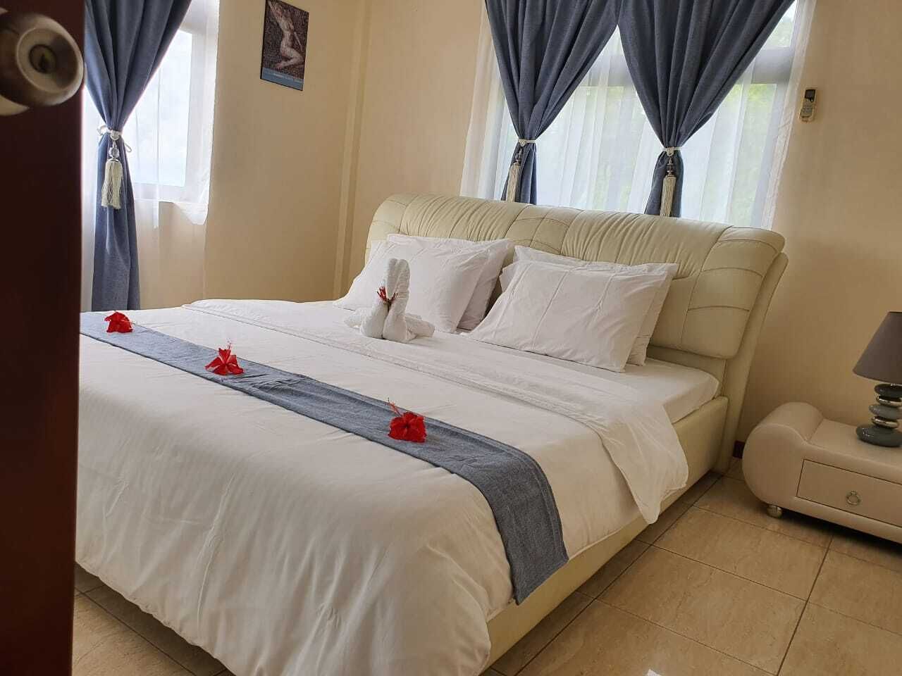 A neatly made bed with white linens and red accents, a bedside table with a lamp, and a window with blue curtains.