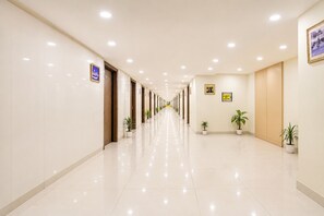 A long, well-lit hallway with potted plants, doors, and signs on the walls. - Hotel D'Capitol- Delhi Airport (New Delhi)