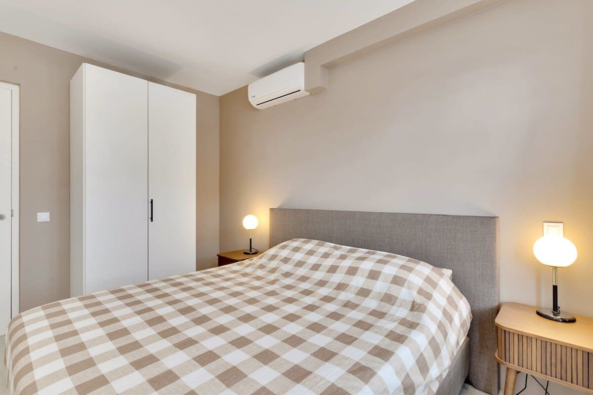 A bedroom with a bed, a wardrobe, a bedside table with a lamp, and an air conditioning unit.