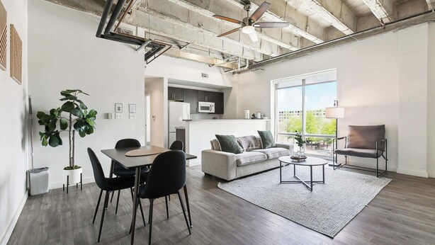 A spacious living area with a dining table, sofa, and armchairs, a kitchen in the background, and a large window providing natural light. - Stay in the Heart of Downtown! (Baton Rouge)