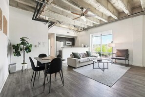 A spacious living area with a dining table, sofa, and armchairs, a kitchen in the background, and a large window providing natural light. - Stay in the Heart of Downtown! (Baton Rouge)