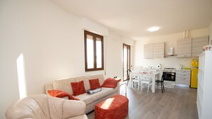 Un salon moderne avec un canapé et un pouf, un coin repas avec une table et des chaises, et une cuisine équipée de meubles et d’appareils électroménagers.