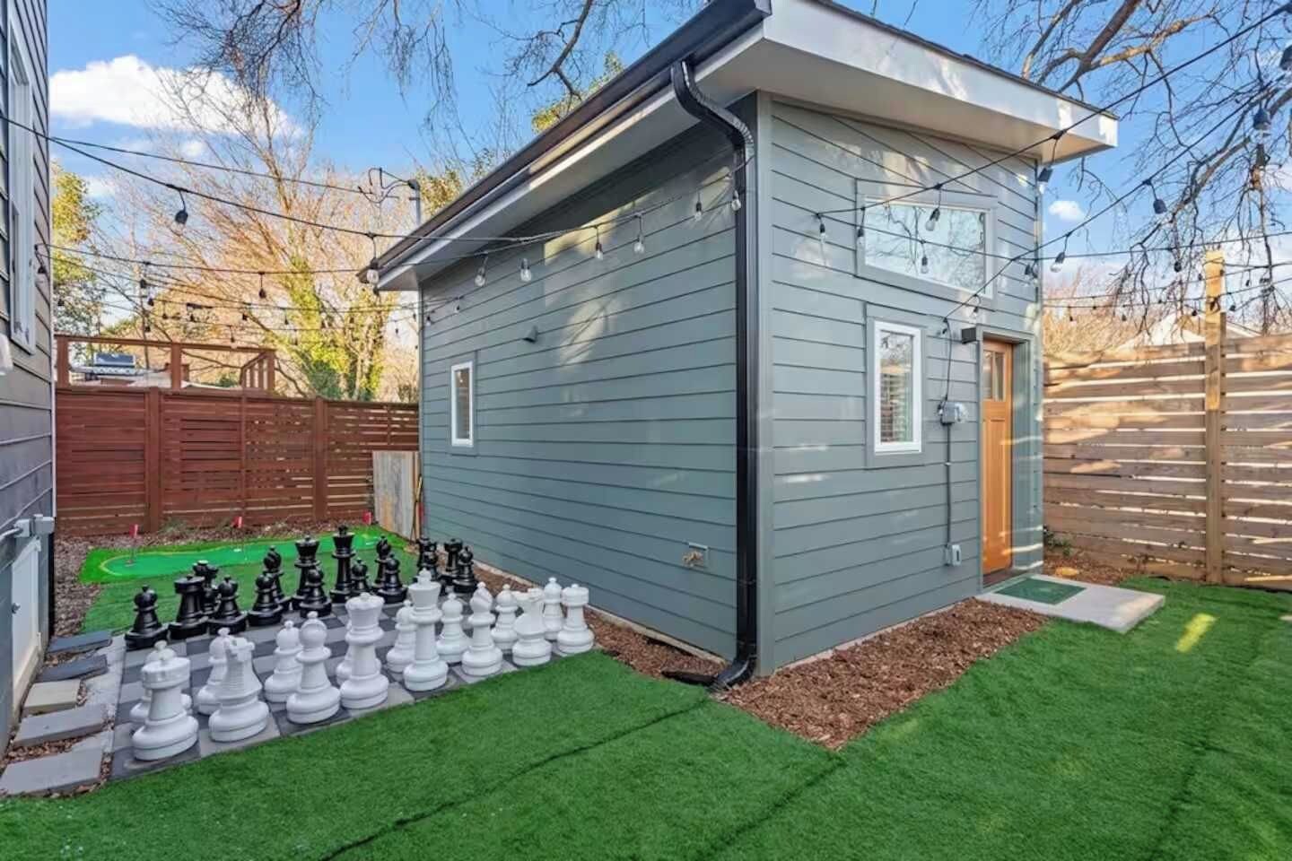 Un cobertizo pequeño con puerta y ventanas, ubicado en un patio con césped artificial y un tablero de ajedrez.