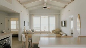 A spacious living area with a high ceiling, a ceiling fan, a large window with a view, a sofa, a coffee table, and a TV mounted on the wall. - The Sundance Cottage (Wrightsville Beach)