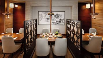 A dining area with a wooden table set for two, surrounded by white chairs, with a traditional Chinese painting on the wall and a decorative screen in the background.