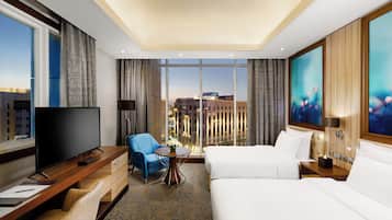A hotel room with a large flat-screen TV, a sofa, a chair, a small table with a vase, and a large window with a city view.