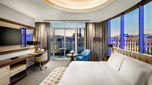 A hotel room with a large bed, a desk with a TV, a chair, a small table with a vase, and a balcony with a city view. - Millennium Place Doha (Doha)