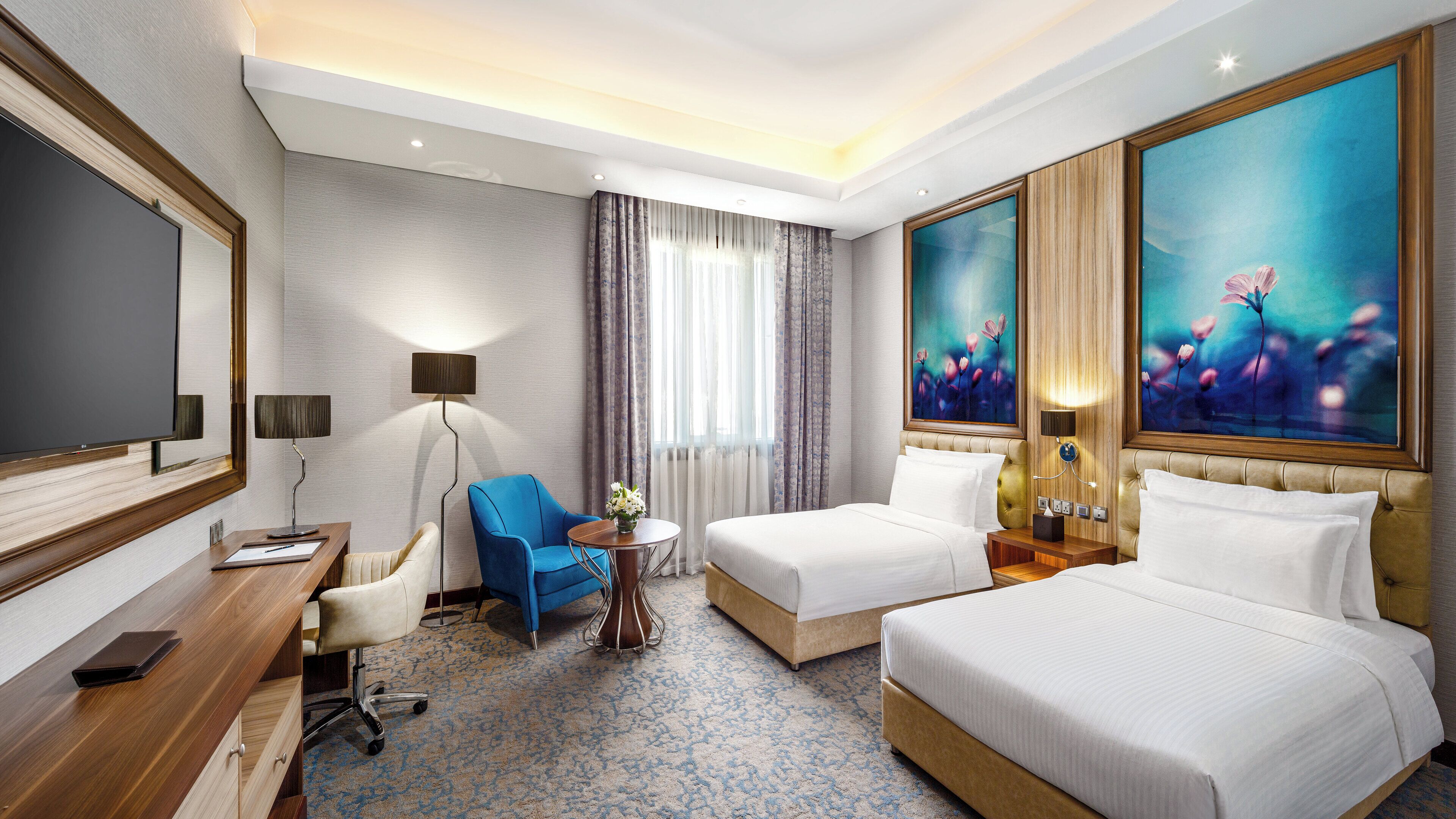 Habitación de hotel con una cama grande, una cama más pequeña, un escritorio con una silla, un sillón azul, una lámpara y un cuadro en la pared.