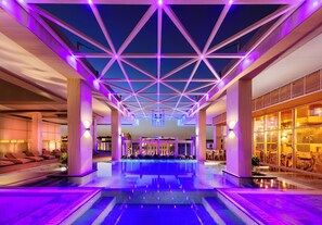 Una piscina interior moderna con iluminación de techo geométrica, áreas de descanso y un restaurante con paredes de vidrio.