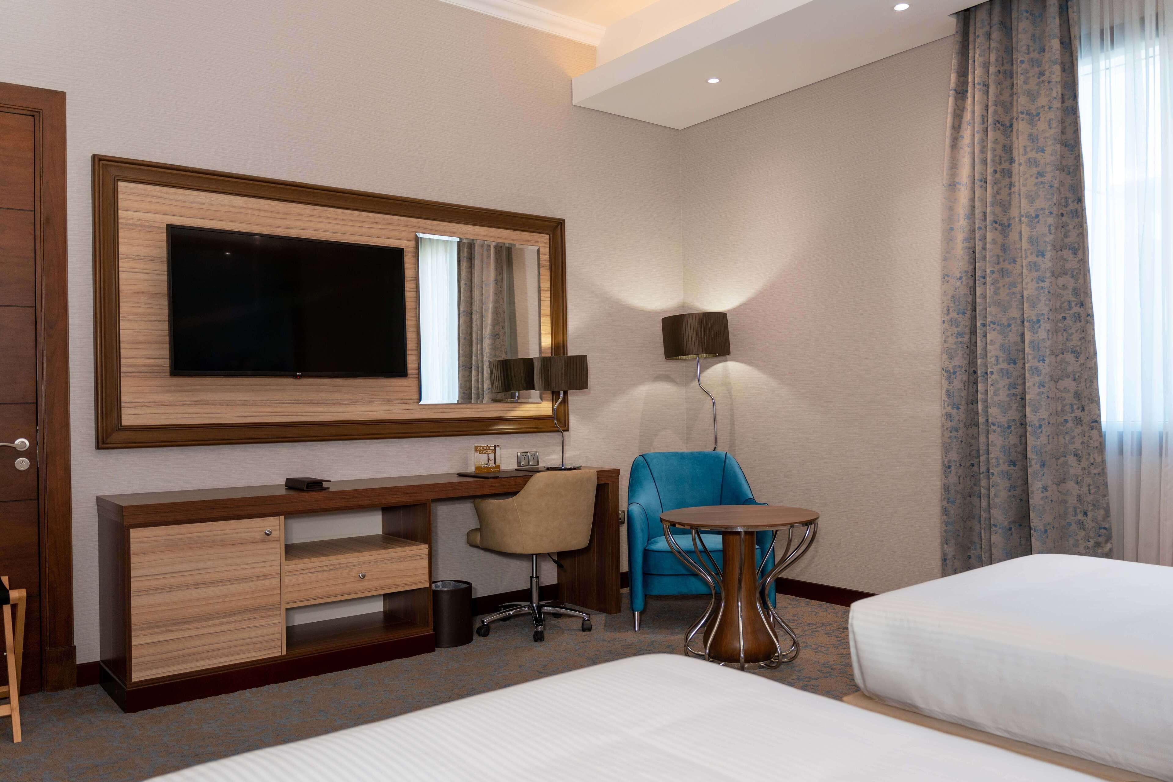 A hotel room with a bed, a desk with a chair, a TV, a lamp, and a small table.