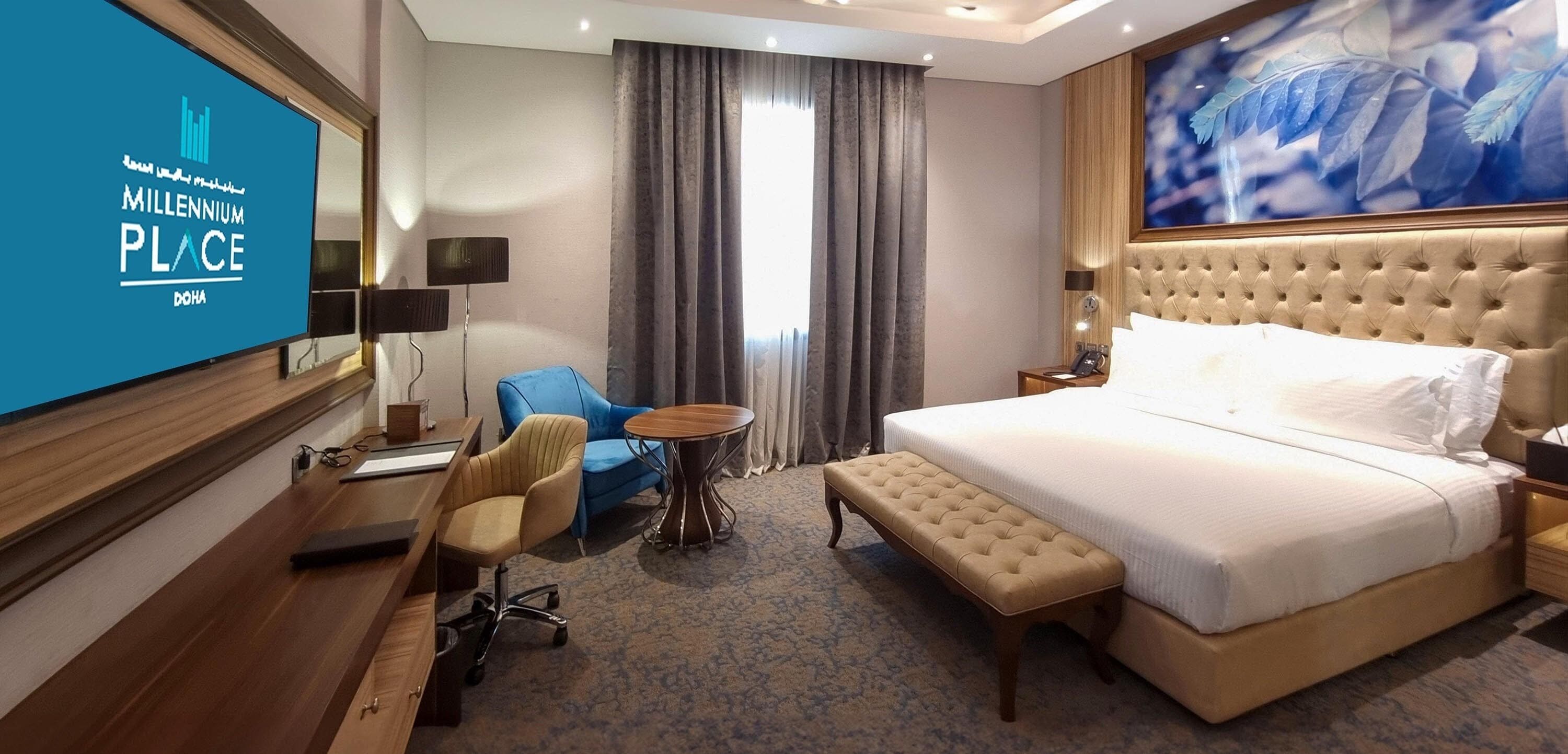 A hotel room with a large bed, a desk, a chair, a small round table, and a TV screen displaying 'Millennium Place Doha'.