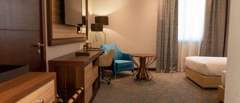 Habitación de hotel con un escritorio de madera, un sillón azul, una mesa redonda, una cama y un espejo grande.
