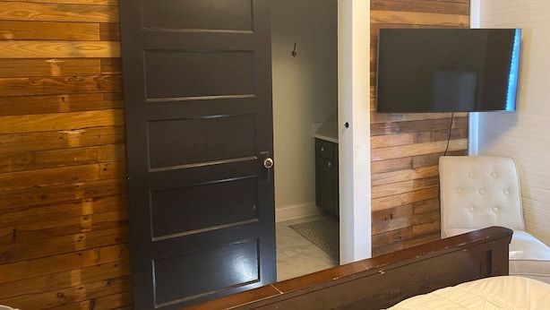 Een slaapkamer met een wand van houten latten, een schuifdeur, een bed, een witte stoel en een aan de muur bevestigde televisie.