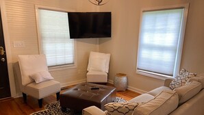 Een woonkamer met een beige bank, twee witte fauteuils, een bruin poefje, een rug met luipaardprint en een flatscreen televisie aan de muur.
