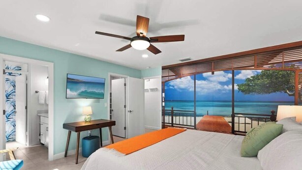 A bedroom with a bed, a desk, a chair, and a view of the ocean through large windows. - 2 Homes, 2 Pools, 2 Game Rooms, Sleeps 21 | IMG (Bradenton)