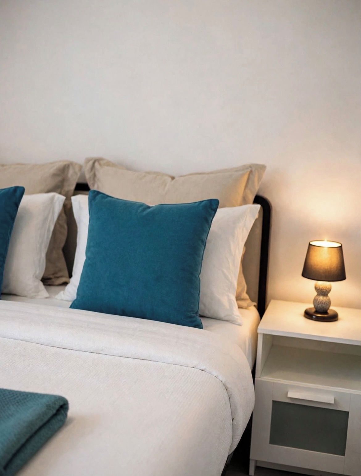A neatly made bed with a white and teal color scheme, a bedside table with a lamp, and a white nightstand.