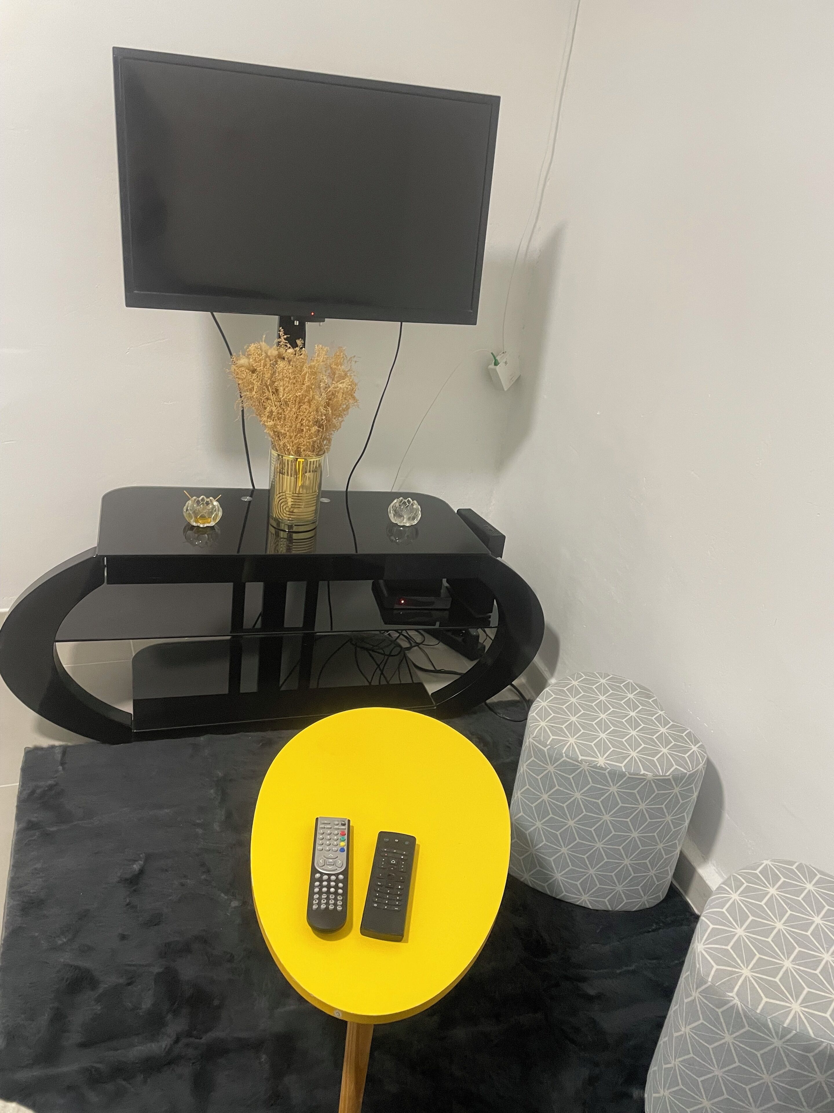 En stue med et fladskærms-tv monteret på et sort stativ, et gult bord med to fjernbetjeninger og en dekorativ vase med tørrede blomster.