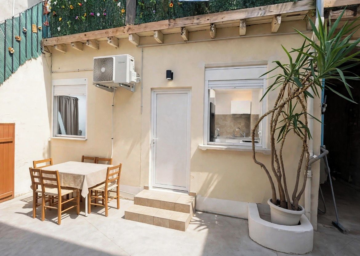 A small outdoor patio with a table and chairs, a potted plant, and a door leading to a covered area.