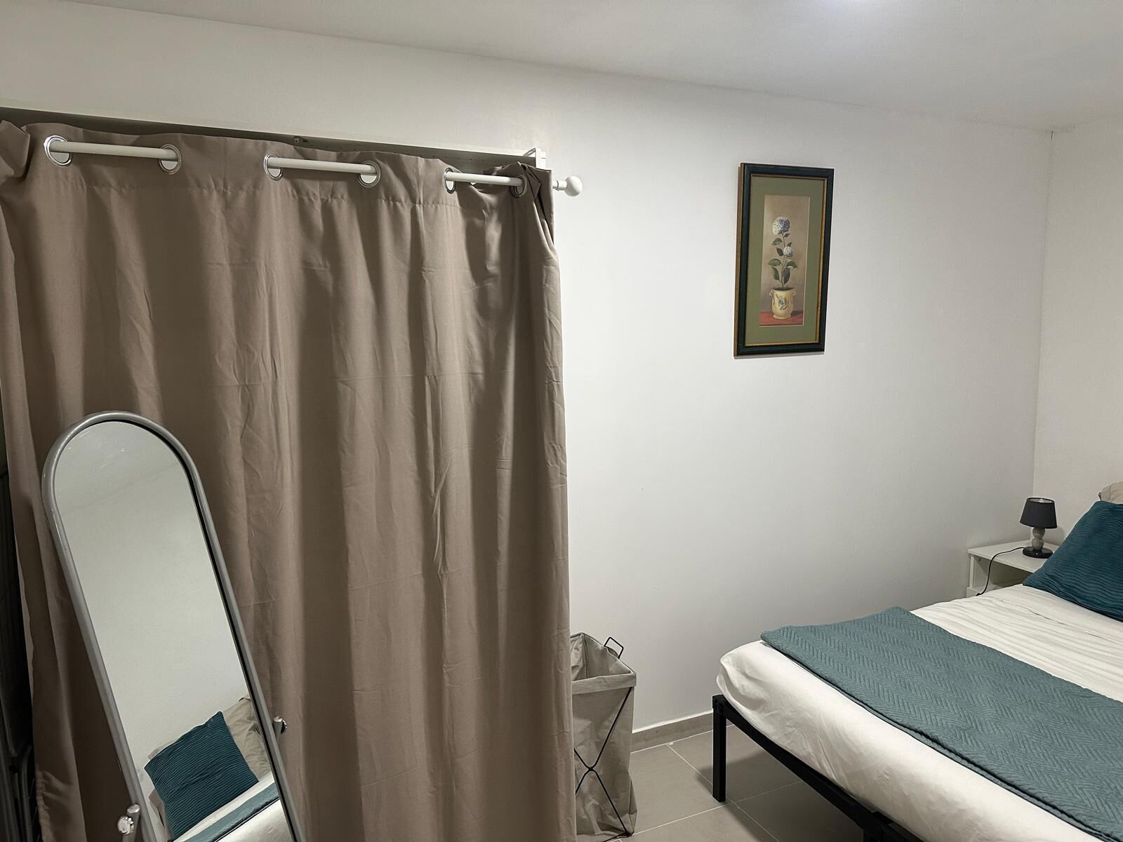 A bedroom with a bed, a bedside table with a lamp, a trash bin, a framed picture on the wall, and a bathroom with a shower curtain and a mirror.