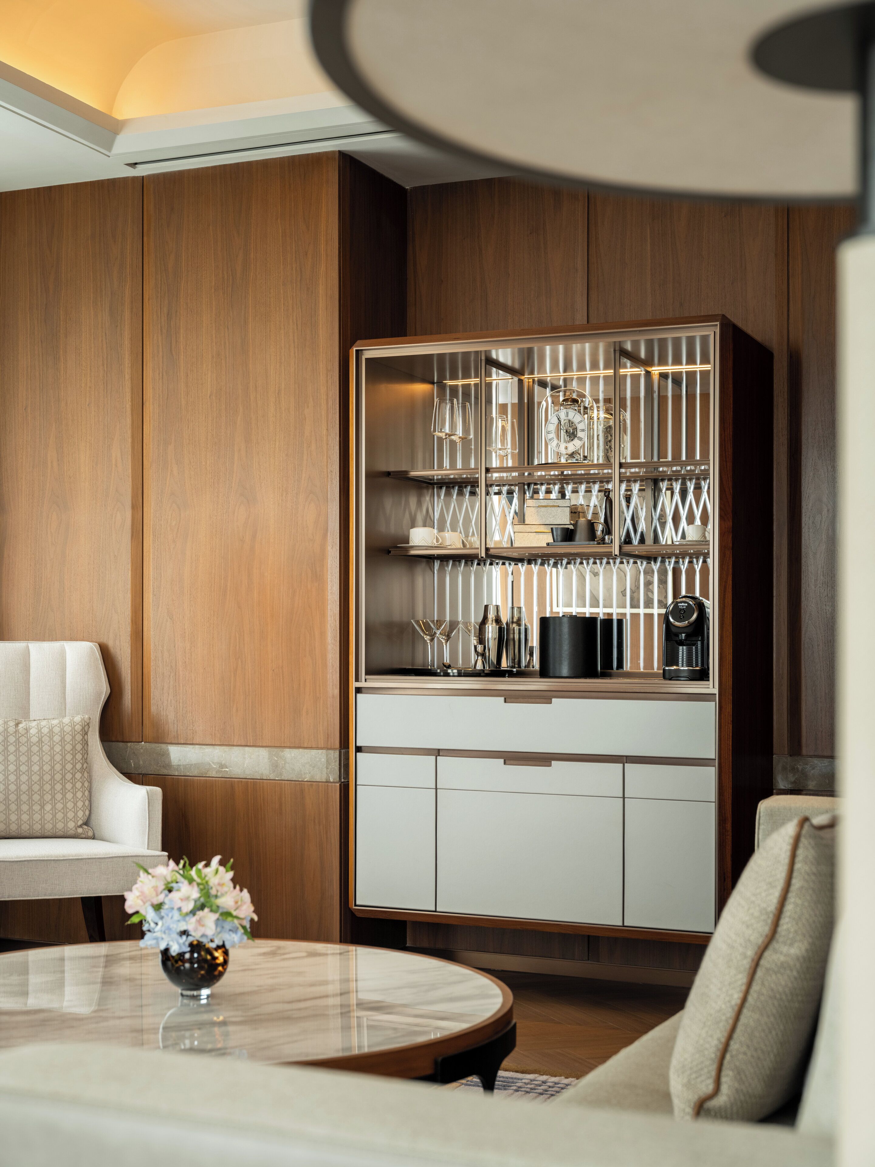 Un salon moderne avec un mur lambrissé en bois, un canapé blanc, une table basse ronde ornée d’un vase de fleurs et un meuble encastré avec des portes en verre, dans lequel sont exposés des ustensiles de cuisine.