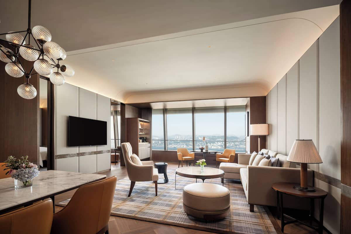 Una habitación de hotel moderna con un gran ventanal, un televisor de pantalla plana, un comedor con mesa de mármol y una zona de estar con sofás y una mesita de café.