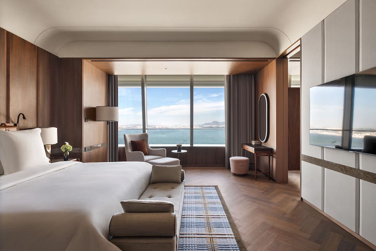 Habitación de hotel moderna con una cama grande, un área de descanso con una silla y un reposapiés, una mesita con lámpara, televisión y vistas al mar.