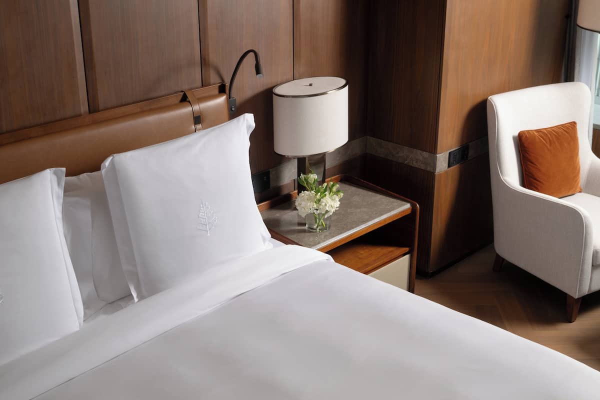 Habitación de hotel con una cama grande, una mesita de noche con lámpara, una silla y un pequeño florero con flores.