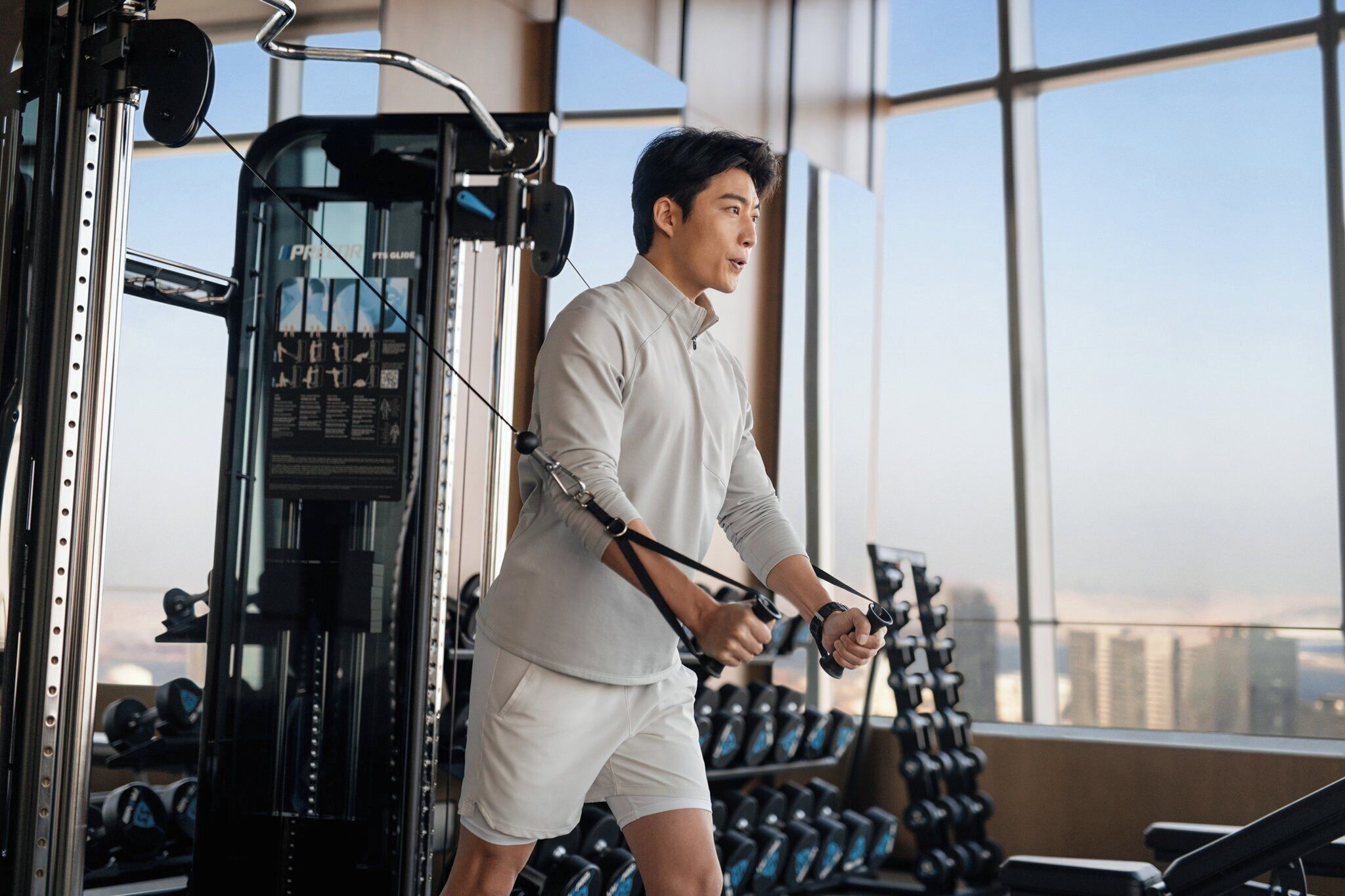 Un gimnasio con equipo de ejercicio y vistas a la ciudad a través de amplias ventanas.