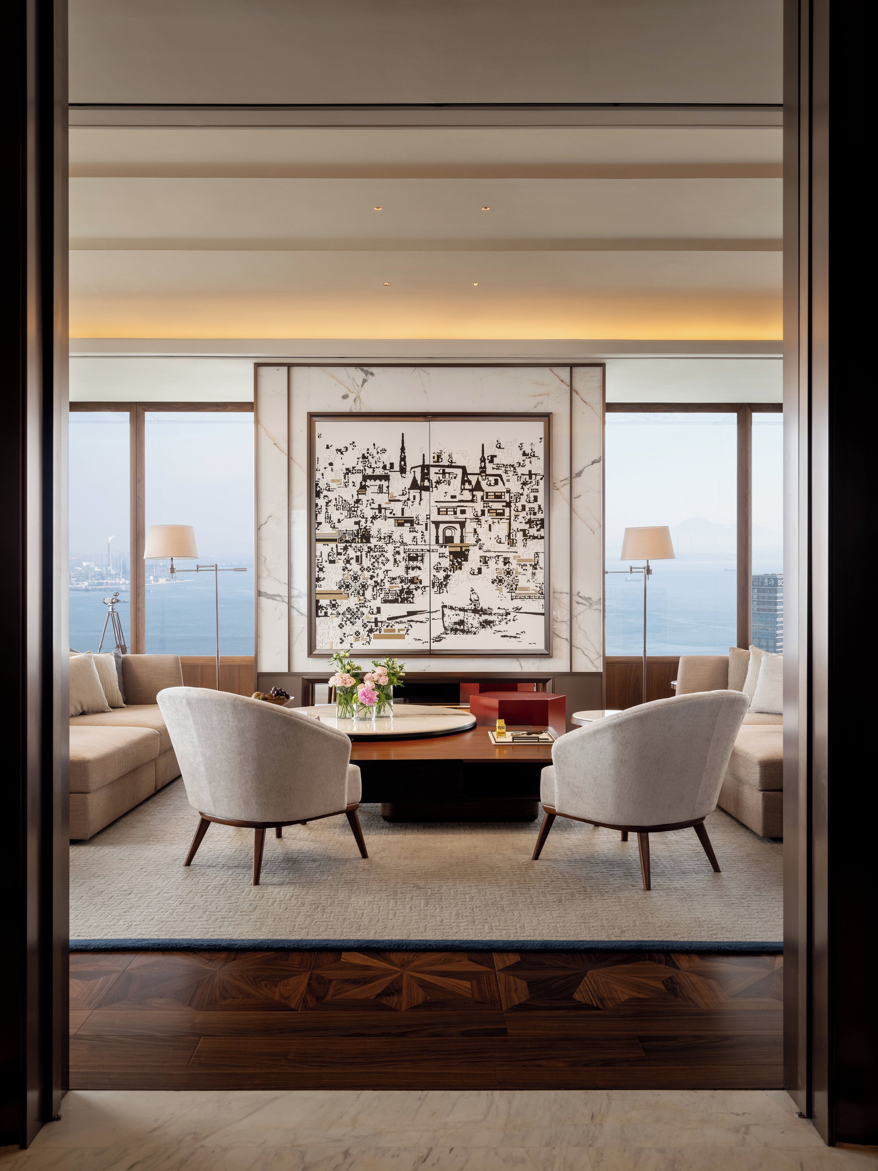 Una sala de estar moderna con una gran obra de arte que representa un paisaje urbano, dos sillones, una mesa de centro y vistas de la ciudad a través de amplios ventanales.